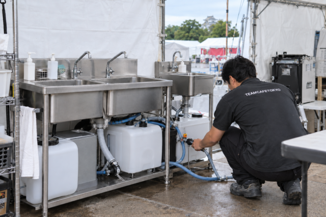 仮設厨房のシンク・給排水タンクと接続部を点検するスタッフ